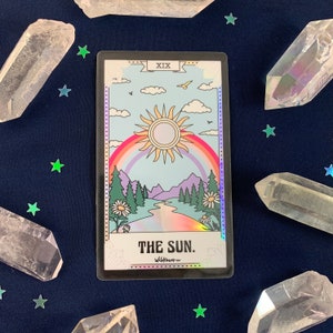 Puede incluir: La carta del tarot El Sol con un fondo negro y un acabado hologr&aacute;fico. La carta presenta una ilustraci&oacute;n colorida de un sol, un arco&iacute;ris, monta&ntilde;as y un r&iacute;o. El n&uacute;mero de la carta XIX est&aacute; en la esquina superior izquierda y el texto "THE SUN." est&aacute; en el centro inferior.