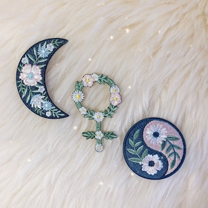 May include: Three embroidered iron-on patches with floral designs. The patches are a crescent moon, a yin-yang symbol, and a female symbol with a flower crown.
