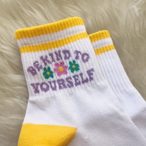 May include: White ankle socks with yellow stripes and a purple message that says "Be Kind To Yourself" with three colorful flowers.