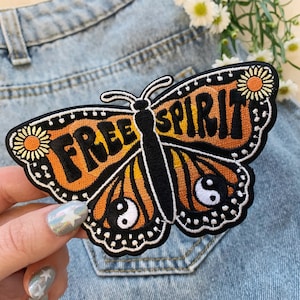 May include: An embroidered iron-on patch featuring a butterfly with orange and black wings. The butterfly has the words "FREE SPIRIT" written on its wings and a yin-yang symbol on each wing.