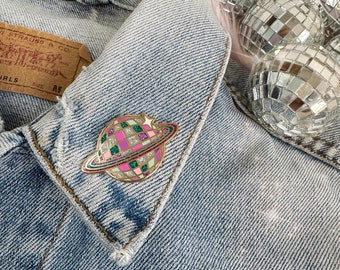 Puede incluir: Un pin de esmalte dorado con un planeta bola de discoteca con cuadrados rosas, verdes y blancos. El planeta tiene un anillo dorado a su alrededor y una pequeña estrella dorada en la parte superior. El pin está en una chaqueta vaquera.