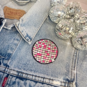 May include: A pink and silver disco ball patch on a blue denim jacket. The patch is surrounded by several silver disco balls.