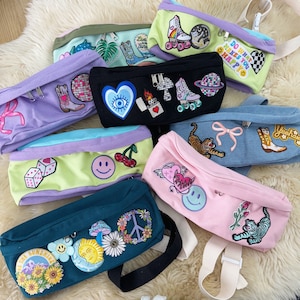 May include: A collection of colorful fanny packs with various embroidered patches. The packs feature designs like roller skates, hearts, smiley faces, and flowers. Each pack has a zipper closure and adjustable strap, with a variety of colors including pink, blue, and green.