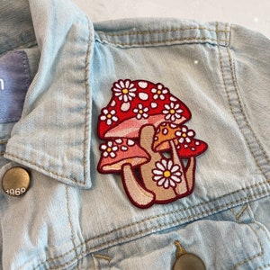 May include: A colorful mushroom patch with red caps, white daisy accents, and tan stems is sewn onto a light blue denim jacket. The patch features a whimsical design, perfect for adding a touch of nature-inspired flair to clothing or accessories.