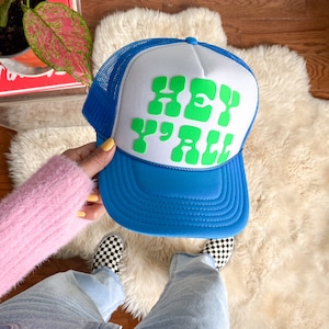 May include: A blue and white trucker hat with a green "Hey Y'all" graphic. The hat has a mesh back and a blue brim.