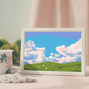 May include: A framed pixel art landscape depicting a blue sky with pink clouds and a green field with sheep. A white mug with floral patterns is placed beside the frame on a pink surface.