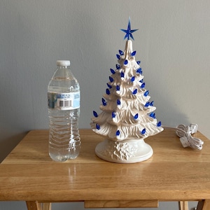 May include: A ceramic Christmas tree with blue lights and a blue star on top. The tree is white and sits on a white base. A water bottle is next to the tree. The tree is approximately 25 cm tall.