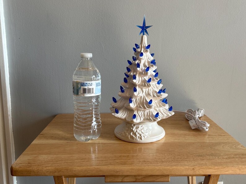 May include: A ceramic Christmas tree with blue lights and a blue star on top. The tree is white and sits on a white base. A water bottle is next to the tree. The tree is approximately 25 cm tall.