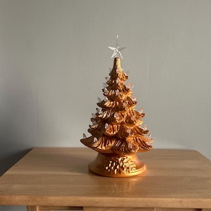 May include: A gold ceramic Christmas tree with clear lights and a star on top. The tree is on a wooden table. The tree is a festive decoration for the holiday season.
