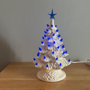 May include: A white ceramic Christmas tree with blue lights and a blue star on top. The tree is lit up and has a classic, vintage design. It is sitting on a wooden surface.