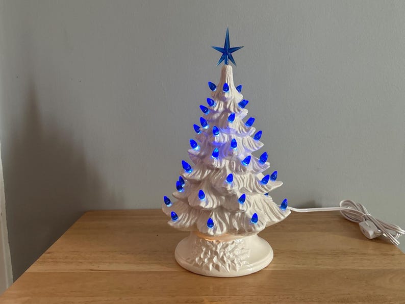 May include: A white ceramic Christmas tree with blue lights and a blue star on top. The tree is lit up and has a classic, vintage design. It is sitting on a wooden surface.