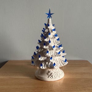 May include: A white ceramic Christmas tree with blue lights and a blue star on top. The tree has a detailed, textured surface and sits on a decorative base. A classic holiday decoration.