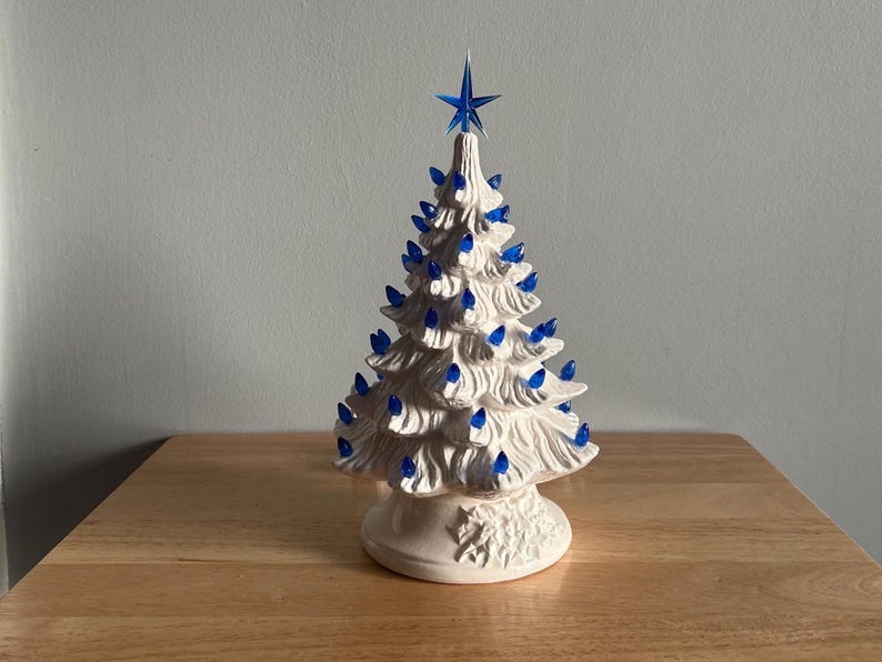 May include: A white ceramic Christmas tree with blue lights and a blue star on top. The tree has a detailed, textured surface and sits on a decorative base. A classic holiday decoration.