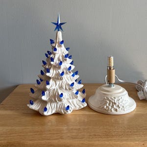 May include: A white ceramic Christmas tree with blue lights and a blue star on top. The tree has a textured surface. A separate base with a light bulb and cord is next to the tree.