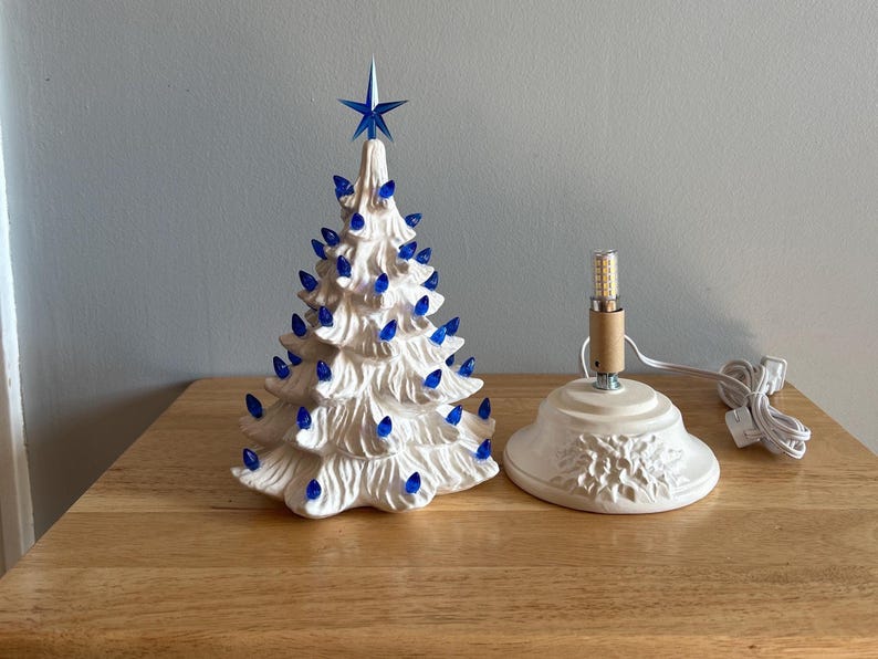 May include: A white ceramic Christmas tree with blue lights and a blue star on top. The tree has a textured surface. A separate base with a light bulb and cord is next to the tree.