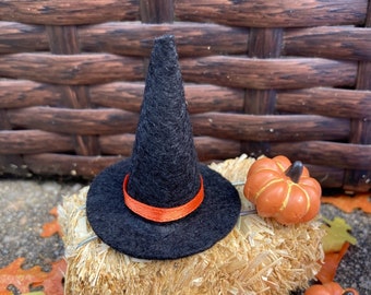 May include: A black felt witch hat with an orange ribbon band sits atop a rectangular straw bale. Two small orange pumpkins with brown stems are positioned next to the straw bale, surrounded by scattered orange and yellow artificial leaves.