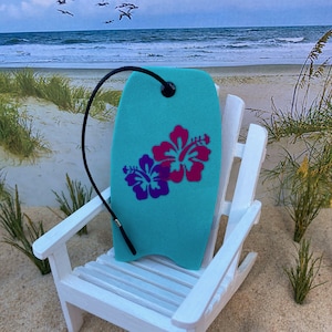 May include: A turquoise surfboard-shaped car air freshener with a pink and purple hibiscus flower design. The air freshener is hanging from a white wooden chair on a sandy beach.