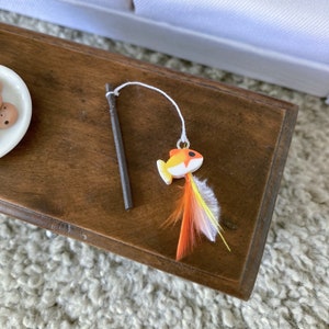 May include: A wooden toy fishing rod with a white string and a small orange and white fish with feathers attached.