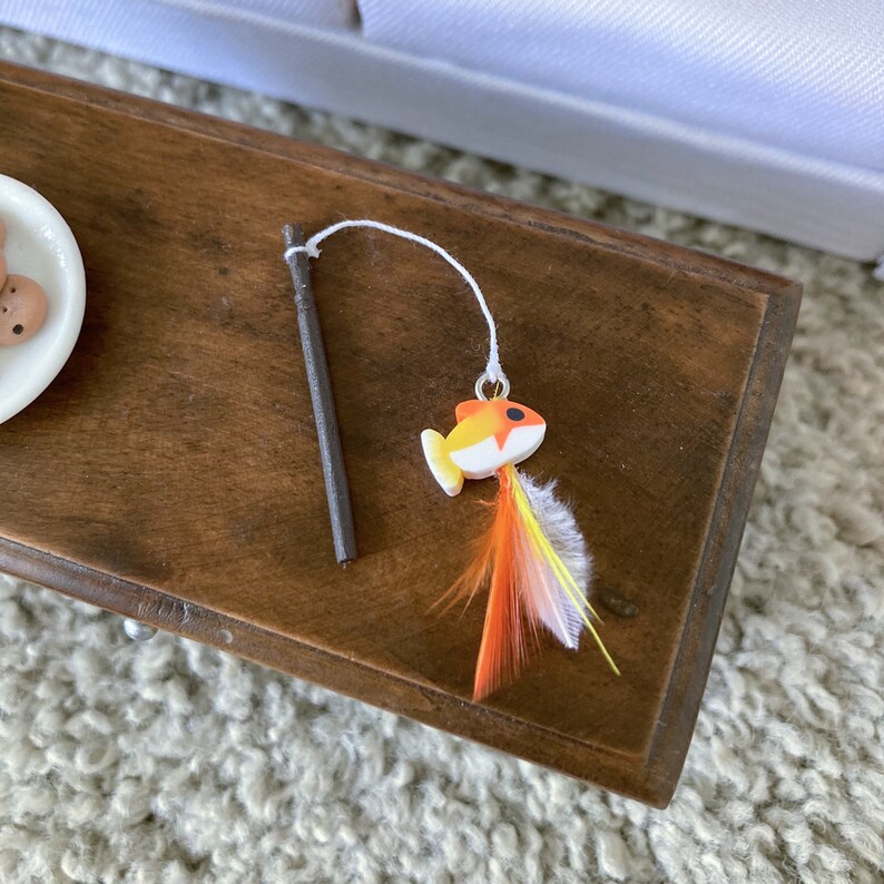 May include: A wooden toy fishing rod with a white string and a small orange and white fish with feathers attached.