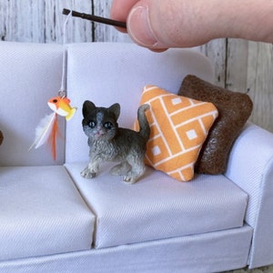 May include: A small gray and white kitten figurine playing with a toy fish on a white couch with orange and brown pillows.