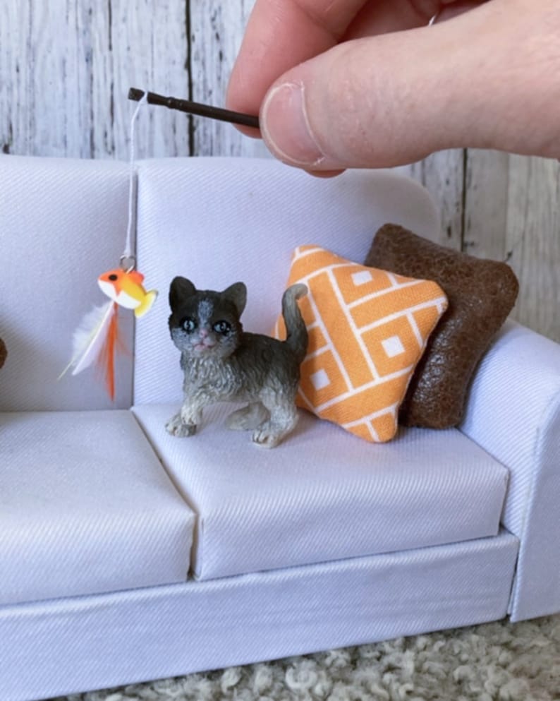 May include: A small gray and white kitten figurine playing with a toy fish on a white couch with orange and brown pillows.
