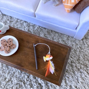 May include: A wooden toy fishing rod with a colorful fish attached to the end. The fish has orange, yellow, and white feathers. The rod is on a wooden table with a plate of chocolate chip cookies.