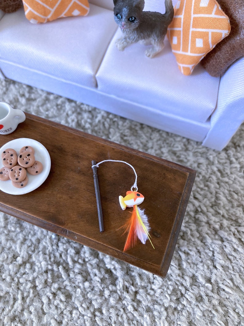 May include: A wooden toy fishing rod with a colorful fish attached to the end. The fish has orange, yellow, and white feathers. The rod is on a wooden table with a plate of chocolate chip cookies.