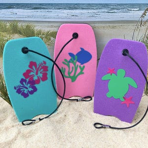 May include: Three foam bodyboards in teal, pink, and purple. The teal board features pink and purple hibiscus flowers. The pink board displays a blue fish and green coral. The purple board shows a green sea turtle and pink starfish. Each has a black leash.
