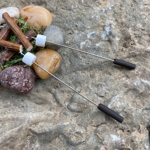 May include: Two silver skewers with white marshmallow roasts and black handles, set on a rock with small logs and stones.