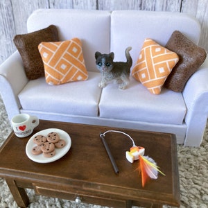 May include: A white couch with brown and orange patterned pillows. A small gray and white kitten is sitting on the couch. A wooden coffee table with a plate of chocolate chip cookies, a small mug, and a toy fish on a string.