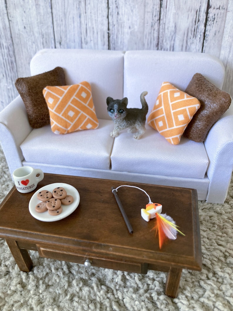 May include: A white couch with brown and orange patterned pillows. A small gray and white kitten is sitting on the couch. A wooden coffee table with a plate of chocolate chip cookies, a small mug, and a toy fish on a string.