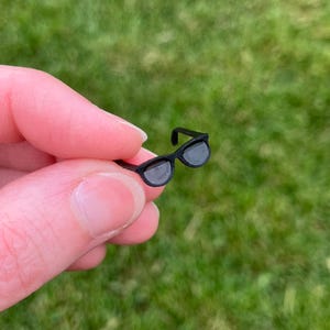 May include: Miniature black sunglasses with gray lenses. The small, round-framed glasses are held against a blurred green background. The sunglasses are a small accessory.