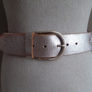 May include: A metallic silver belt with a textured finish and a rounded silver-tone metal buckle. The belt features a pointed tip, a single belt loop, and a series of punched holes for adjustment, secured around a neutral-colored mannequin torso.