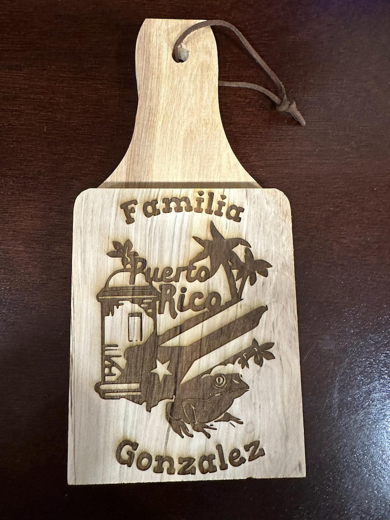 May include: A wooden cutting board with a brown leather strap handle. The board is engraved with the words "Familia", "Puerto Rico", and "Gonzalez". The design also includes a palm tree, a building, and a frog.