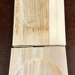 May include: A wooden hinged box with a round cutout in the bottom. The box is made of light wood and has a natural finish.