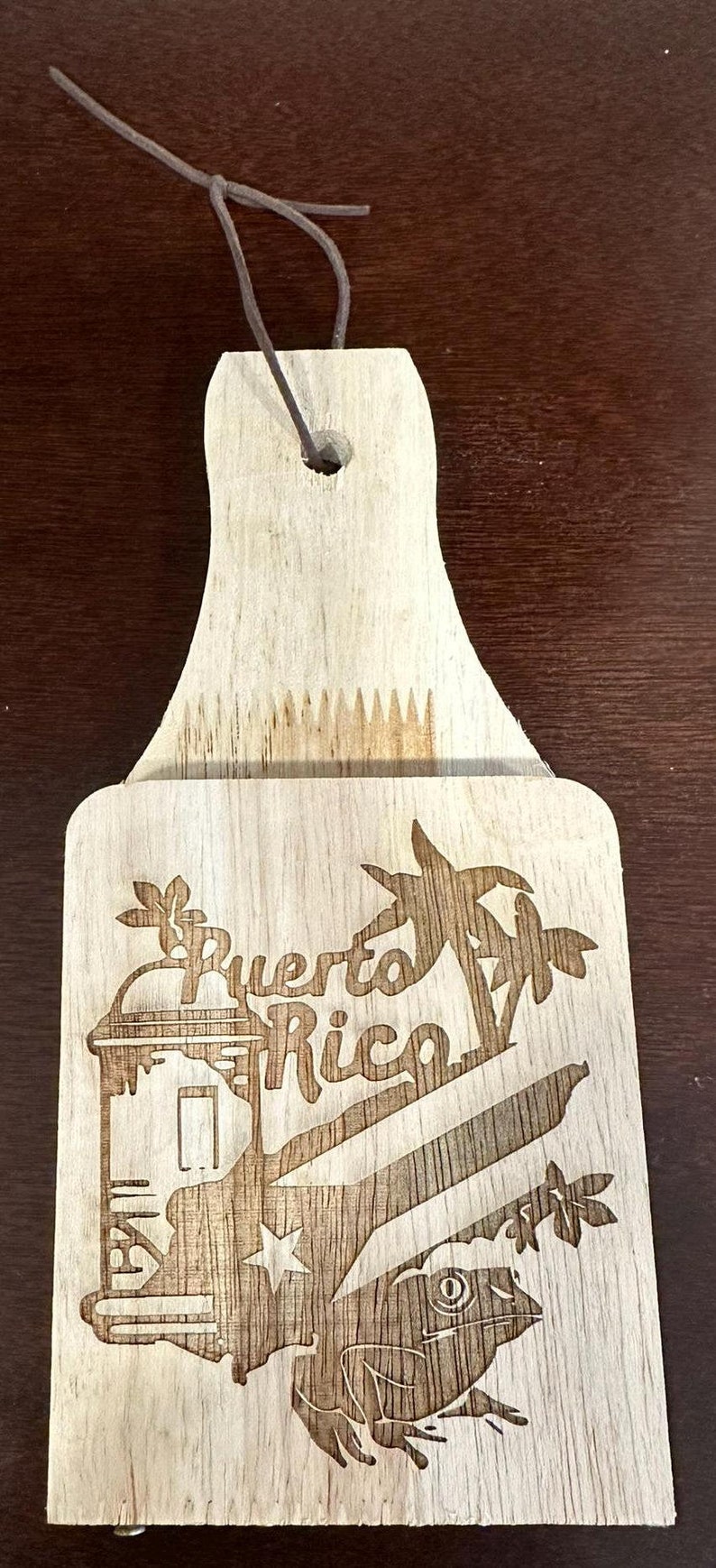 May include: Wooden cutting board with a brown wood grain finish. The board has a hanging hole at the top and a cutout for holding a knife. The board is decorated with a laser-engraved design of a palm tree, a frog, and the words "Puerto Rico".