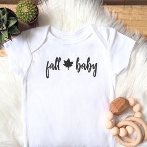 May include: White baby onesie with black text that reads "fall baby" with a black maple leaf between the words.