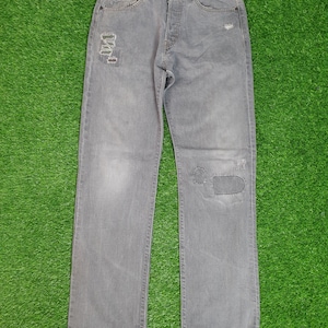 Op de afbeelding: Grijze spijkerbroek met distressed details en patches. De spijkerbroek heeft meerdere gaten en scheuren, en een patch is over een van de gaten genaaid.