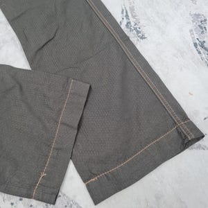 May include: Close-up of olive green pants with a textured pattern. The pants have a straight leg design and are stitched with light brown thread. The fabric appears to be a durable material, suitable for casual wear.