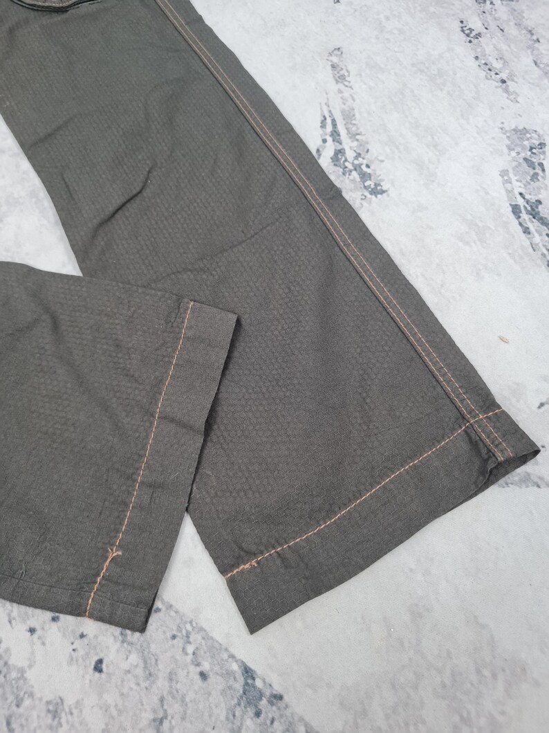 May include: Close-up of olive green pants with a textured pattern. The pants have a straight leg design and are stitched with light brown thread. The fabric appears to be a durable material, suitable for casual wear.