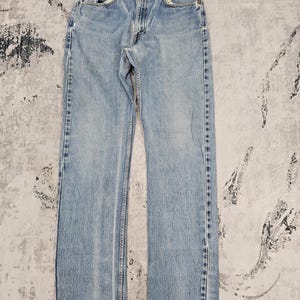 May include: Light blue denim jeans with a classic five-pocket design. The jeans have a straight leg cut and a faded wash, with visible wear and tear details. The jeans are laid flat on a patterned surface.