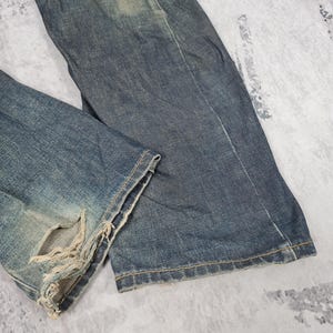 Op de afbeelding: Een paar blauwe denim jeans met distressed details en rafelige randen. De jeans heeft een vervaagde en versleten look, wat hem een vintage uitstraling geeft.