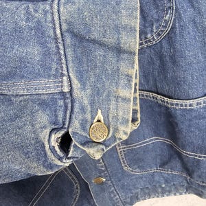 Puede incluir: Primer plano de una chaqueta vaquera azul con cierre de bot&oacute;n y un solo bolsillo en el lado izquierdo. El bot&oacute;n tiene la palabra "SEARS" grabada.