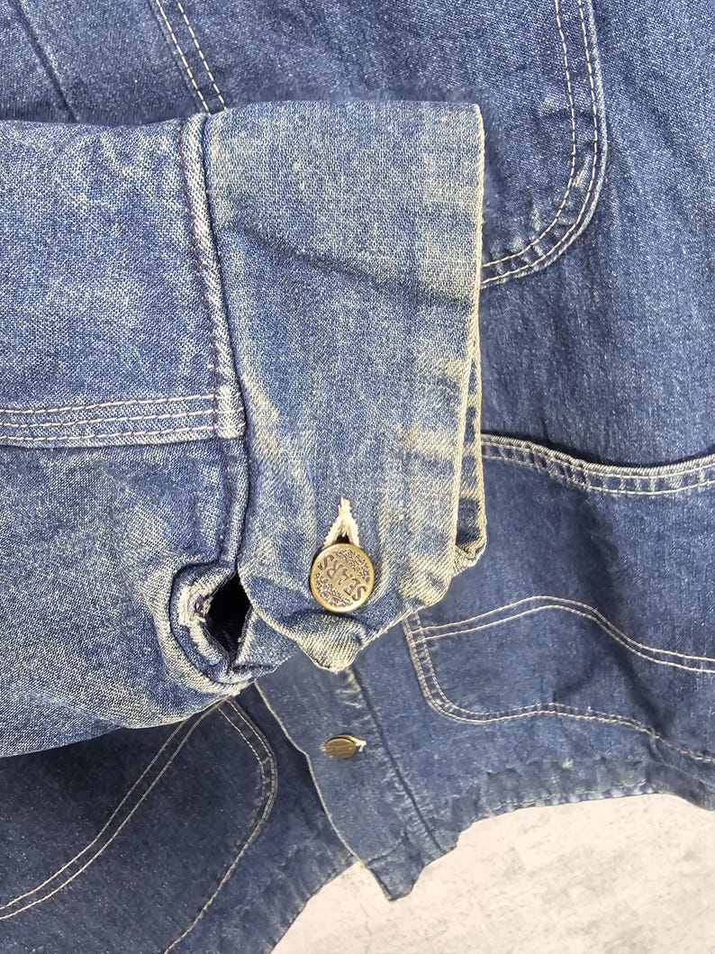 Puede incluir: Primer plano de una chaqueta vaquera azul con cierre de bot&oacute;n y un solo bolsillo en el lado izquierdo. El bot&oacute;n tiene la palabra "SEARS" grabada.
