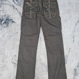 May include: Dark olive-green cargo pants with multiple pockets and decorative stitching. The pants feature large, flap-covered pockets on the back and sides, with adjustable straps and metal hardware. The stitching is a contrasting orange color.