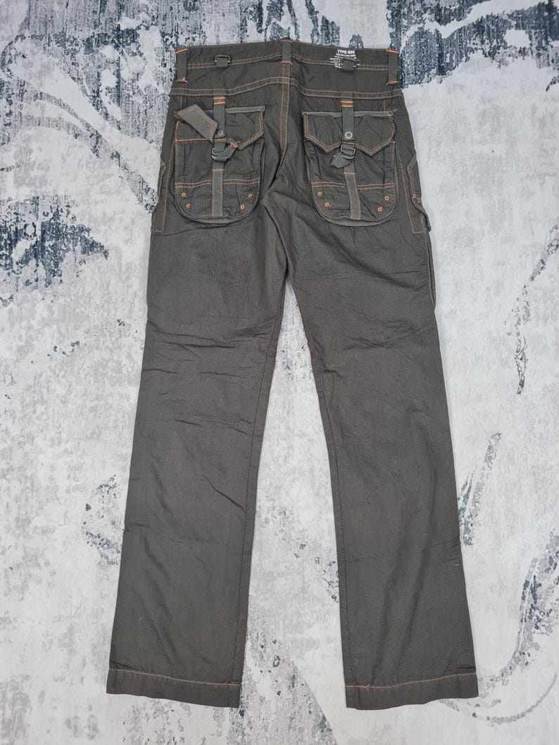 May include: Dark olive-green cargo pants with multiple pockets and decorative stitching. The pants feature large, flap-covered pockets on the back and sides, with adjustable straps and metal hardware. The stitching is a contrasting orange color.