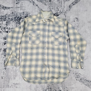 Pode incluir: Uma camisa de manga comprida com botões e um padrão xadrez em azul e creme. A camisa inclui um colarinho pontiagudo, dois bolsos no peito com fechos de pressão e botões de pressão na carcela frontal e nos punhos das mangas.