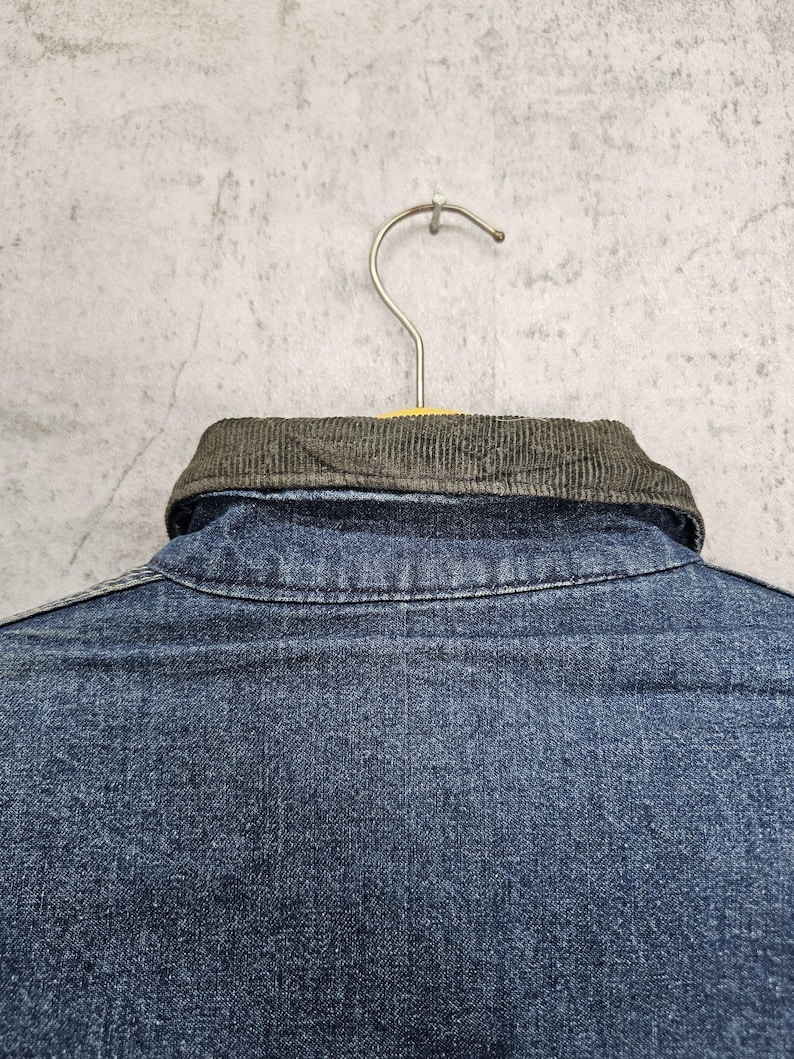 Puede incluir: Una camisa vaquera azul con cuello de pana. La camisa est&aacute; colgada en una percha.