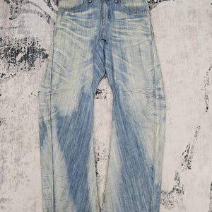 May include: Light blue denim jeans with a faded wash effect. The jeans have a classic five-pocket design, a button and zip closure, and a wide leg. The fabric shows signs of wear, adding to the vintage aesthetic.