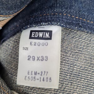 Op de afbeelding: Een wit label met zwarte tekst dat "EDWIN. E2000 SIZE 29 X 33 EEM=277 E505-1406" zegt, bevestigd aan een blauwe spijkerbroek.